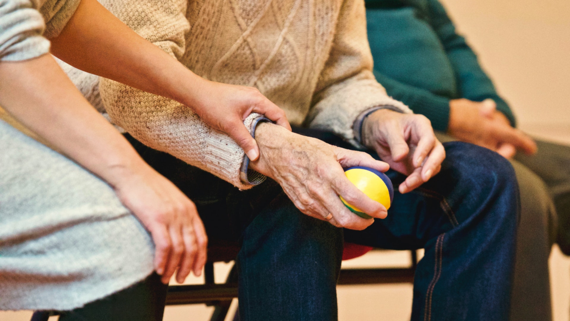 Hands of an elderly person holding a colorful stress ball, assisted by a caregiver, with additional individuals seated nearby, emphasizing support and care in a healthcare or rehabilitation setting related to Medicare fraud awareness.