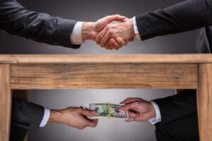 Close-up Of Two Businesspeople Shaking Hand And Taking Bribe Under Wooden Table On Grey Background, representing TRICARE bribery